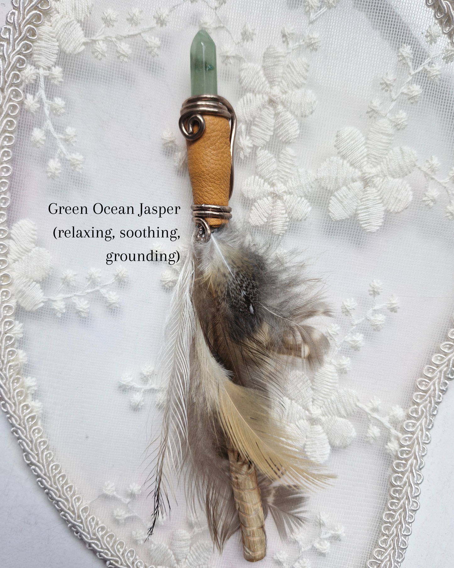 Decorative wand with a green jasper crystal, wooden handle, and feathers on a white lace background.