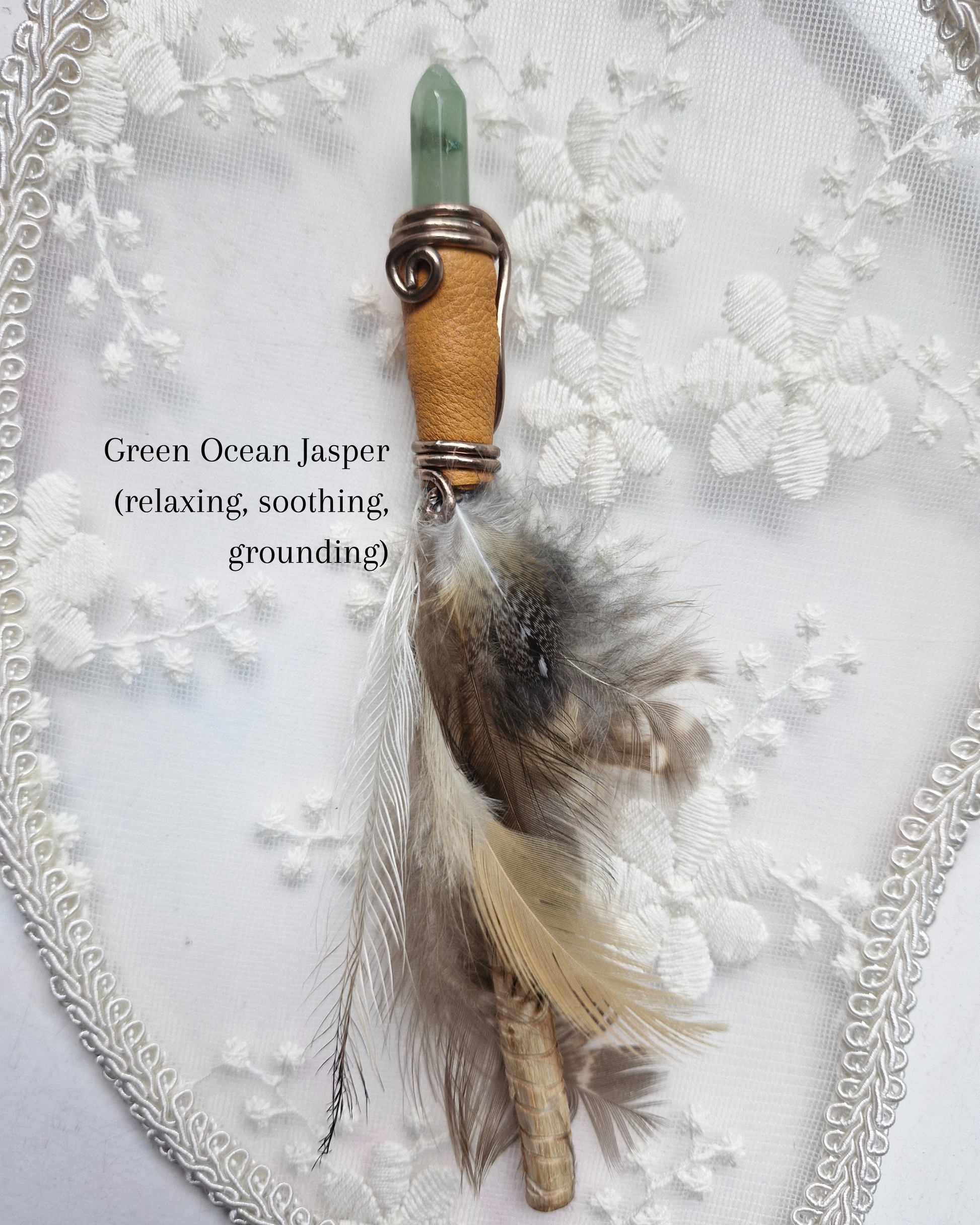 Decorative wand with a green jasper crystal, wooden handle, and feathers on a white lace background.